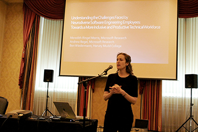 A presenter stands in front of a mic and her powerpoint.