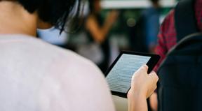 A person holding up a tablet and reading while in a bustling room.