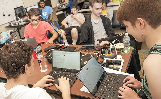 A group of students sit around a table coding on their laptops.