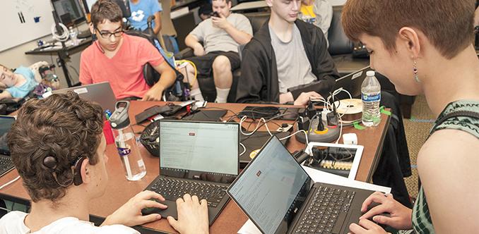 A group of students sit around a table coding on their laptops.