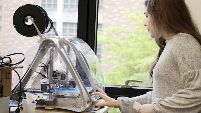 Still showing a woman looking at a 3-D printer.