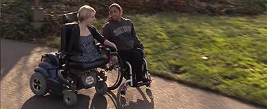 Two students in wheelchairs go down a college path.