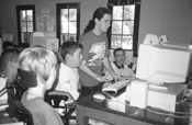 DO-IT Intern, CJ, demaonstrates Internet techniques to a rapt audience at MDA Camp Waskowitz. Picture of campers watching CJ use computer