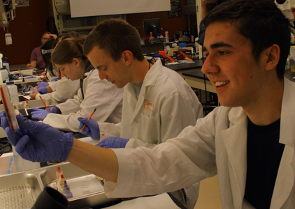 Picture of scholars in lab coats and cloves examining and using lab equipment.