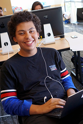 A student using a laptop and headphones smiles at the camera.