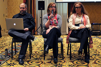 Three panelists sit at the front of the room. One speaks into the microphone. Three panelists sit at the front of the room. One speaks into the microphone.