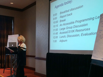 Sheryl Burgstahler stands in front of that day's agenda on the screen at the AccessCS10K CBI.