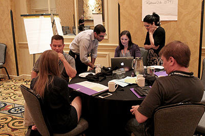 Three participants look at a computer while others speak around the table.