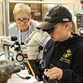 A student looks into a microscope while an educator stands by.