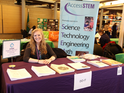 AccessSTEM Team member Jenica sitting behind the DO-IT booth at the 'See It. Be It' Conference. Photo of AccessSTEM Team member Jenica sitting behind the DO-IT booth at the 'See It. Be It' Conference.
