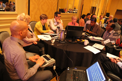 8 professionals sit around a table talking to each other while a captioner writes down what they are saying and displays it on a screen.