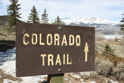 Photo of wood sign marking the Colorado Trail with pine trees and mountains in the background.