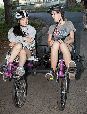 DO-IT Phase I Scholars Chana and Katelyn share a laugh while riding an accessible bike together.