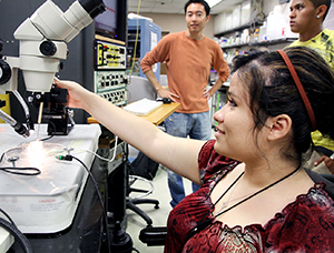 A student in a wheelchair uses a microscope while looking at her faculty advisor.