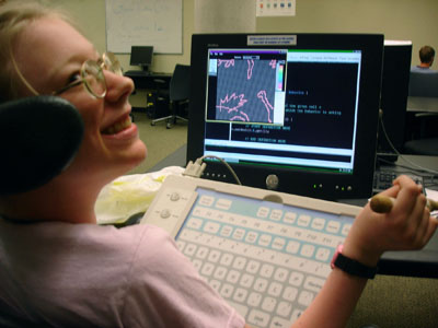 Photo a student in a wheelchair holding a small wooden stick and types into the computer using an adaptive keyboard
