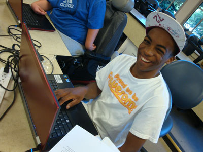 Photo of a male student with cap smiling while working on a laptop