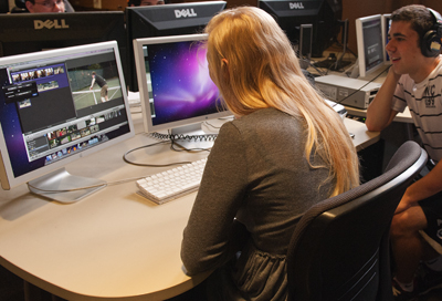 Photo of DO-IT Scholar sitting at a computer with two large displays while DO-IT intern Reese looks on.