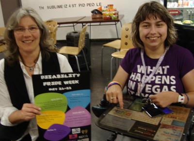 Two people pose for a picture, one is holding a Disability Pride Week poster.