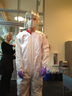 Photo of A student wearing a large white hazmat suit with purple gloves, a paper face mask, and a large plastic face guard stands in the center, while a woman in the back of the shot helps someone else.