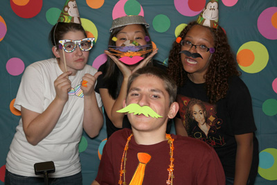 Photo of 2012 Scholars Alicia, Grace, and Aaliyah pose with 2010 Intern Benjy wearing decorated paper glasses and mustaches in front of a polka-dot backdrop.