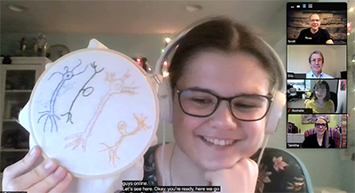 An NNL student shows their science-related cross stitch project to the camera.