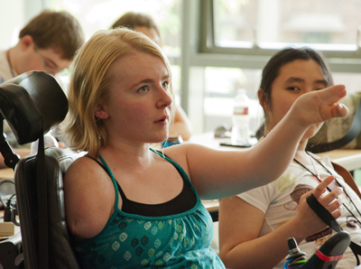 Photo of Phase I DO-IT Scholar Kayla holds up her arm and speaks up in a classroom, with Phase I Scholar Clarissa sitting next to her