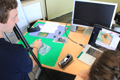 Photo of Phase II DO-IT Scholar Eric is drawing on a piece of paper under a camera, which is also shown on the laptop next to him.