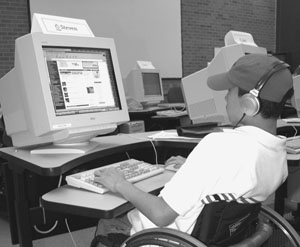 Image of a scholar in a wheelchair sitting in front of a computer wearing headphones