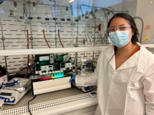 Young woman scientist in labcoat and mask standing next to lab work bench with a row of test tubes containing glowing liquid in each one.