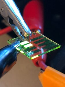 Close up photo of metal clips holding small clear plastic optical chip with glowing sections indicating activity in the areas