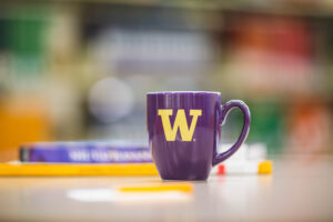A UW-branded purple mug sits on a table in front of a blurred background.