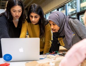 Three women conducting research gather around and look at a laptop.